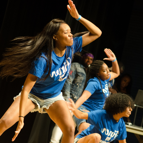 A group of students perform energetic dance moves on stage during the NPHC Stroll Off at Indiana State University, wearing matching blue shirts and shorts.